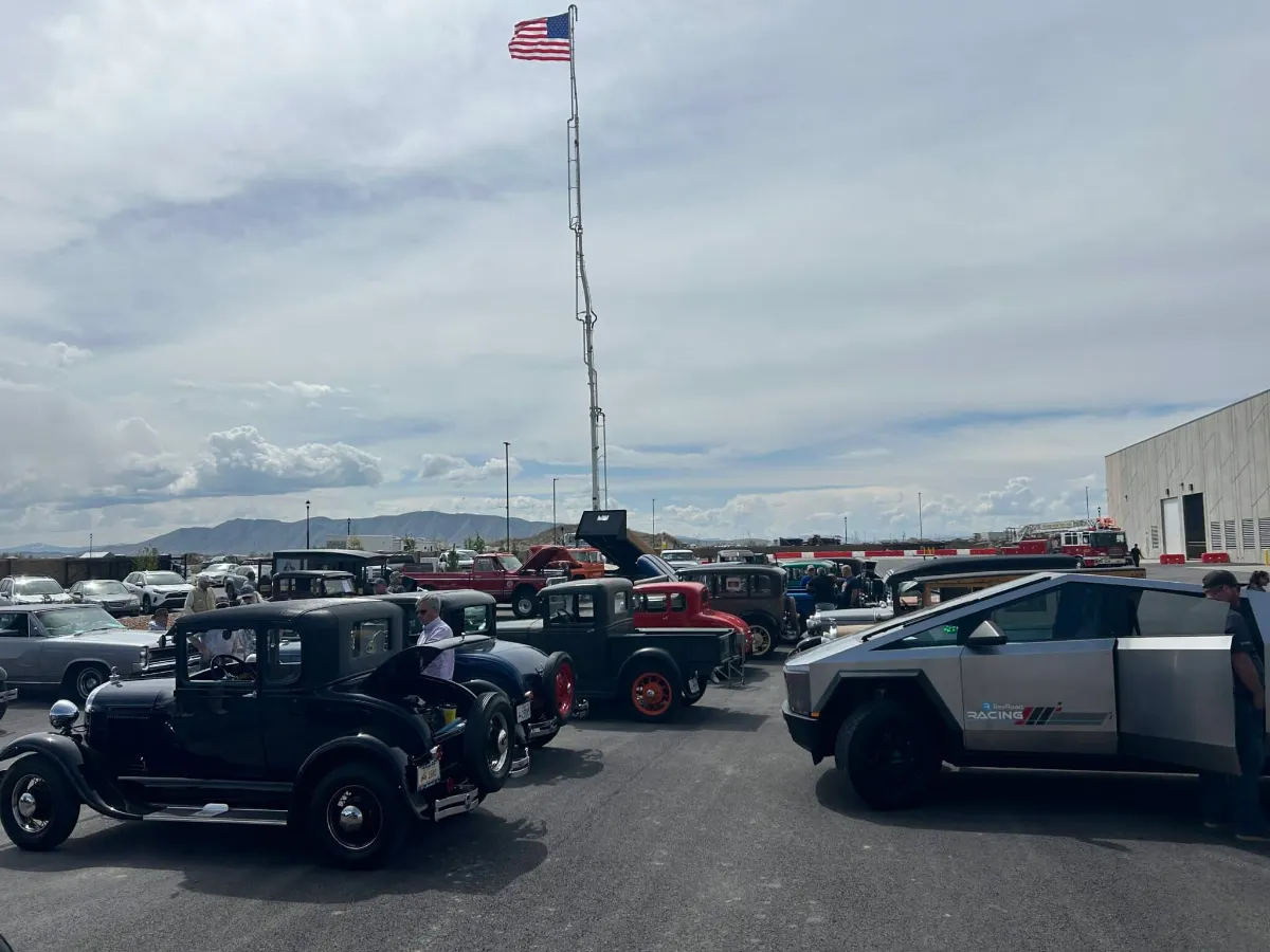 Classic cars and pickup trucks parked under a flagpole at the car show