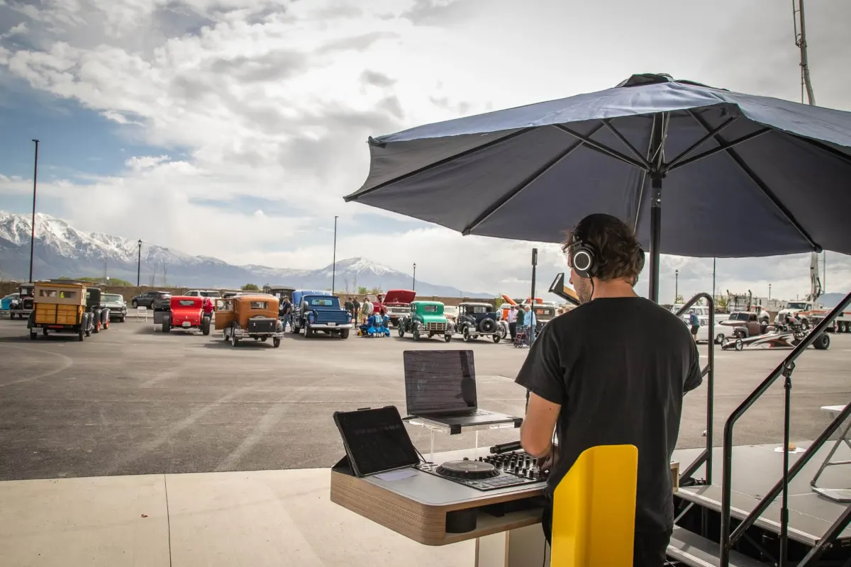 DJ booth set up under a shade umbrella with classic cars lined up across the lot behind it