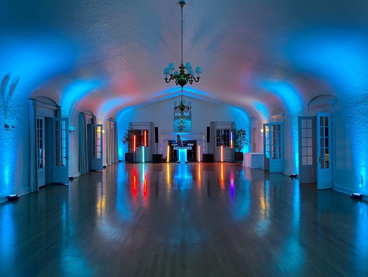 Historic ballroom in blue and teal uplighting with a chandelier and DJ setup glowing at the far end